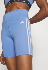 Gros plan sur une personne portant un short de vélo bleu Adidas avec trois bandes blanches sur le côté et un soutien-gorge de sport assorti.