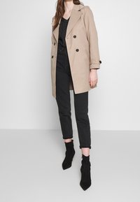 Donna che indossa un cappotto beige a doppio petto, una maglietta nera con pois, jeans neri a vita alta e stivaletti neri con punta, in piedi su uno sfondo unico.