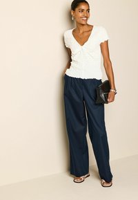 Haut blanc texturé avec un col en V et des manches courtes, associé à un pantalon large taille haute bleu marine et une pochette noire. Des sandales plates complètent le look.