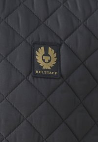 Fekete steppelt anyag, arannyal hímezett logóval, melyen egy sas alakja és az alján a "BELSTAFF" felirat található.