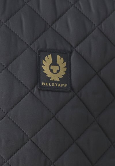 Tissu matelassé noir comportant un patch logo brodé doré avec un design d'aigle et le mot "BELSTAFF" en bas.