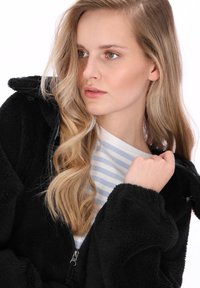 Czarna kurtka polarowa na zamek, o miękkiej fakturze. Nosi się ją na biało-niebieską koszulę w paski, podkreślając warstwowość stroju.