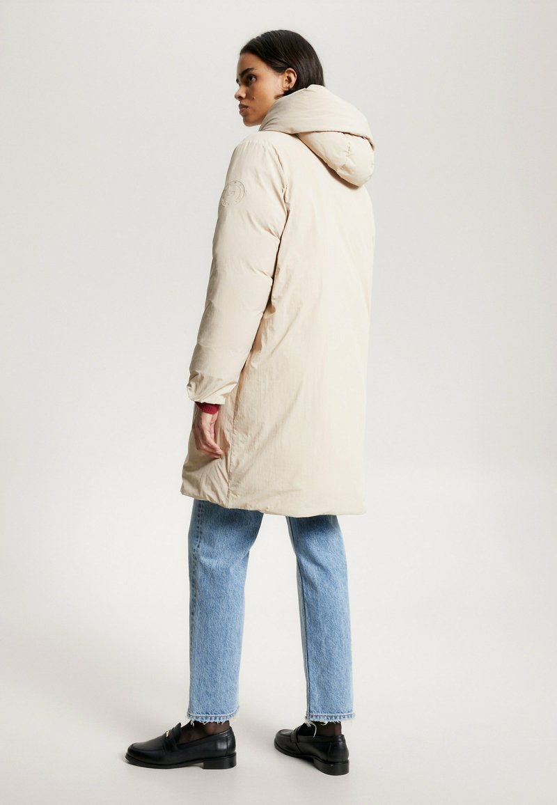 Beige gepolsterter Mantel mit Kapuze, ausgestattet mit einem Logo auf dem Ärmel. Kombiniert mit hellblauen Jeans und schwarzen Loafers, zeigt eine gerade Silhouette.