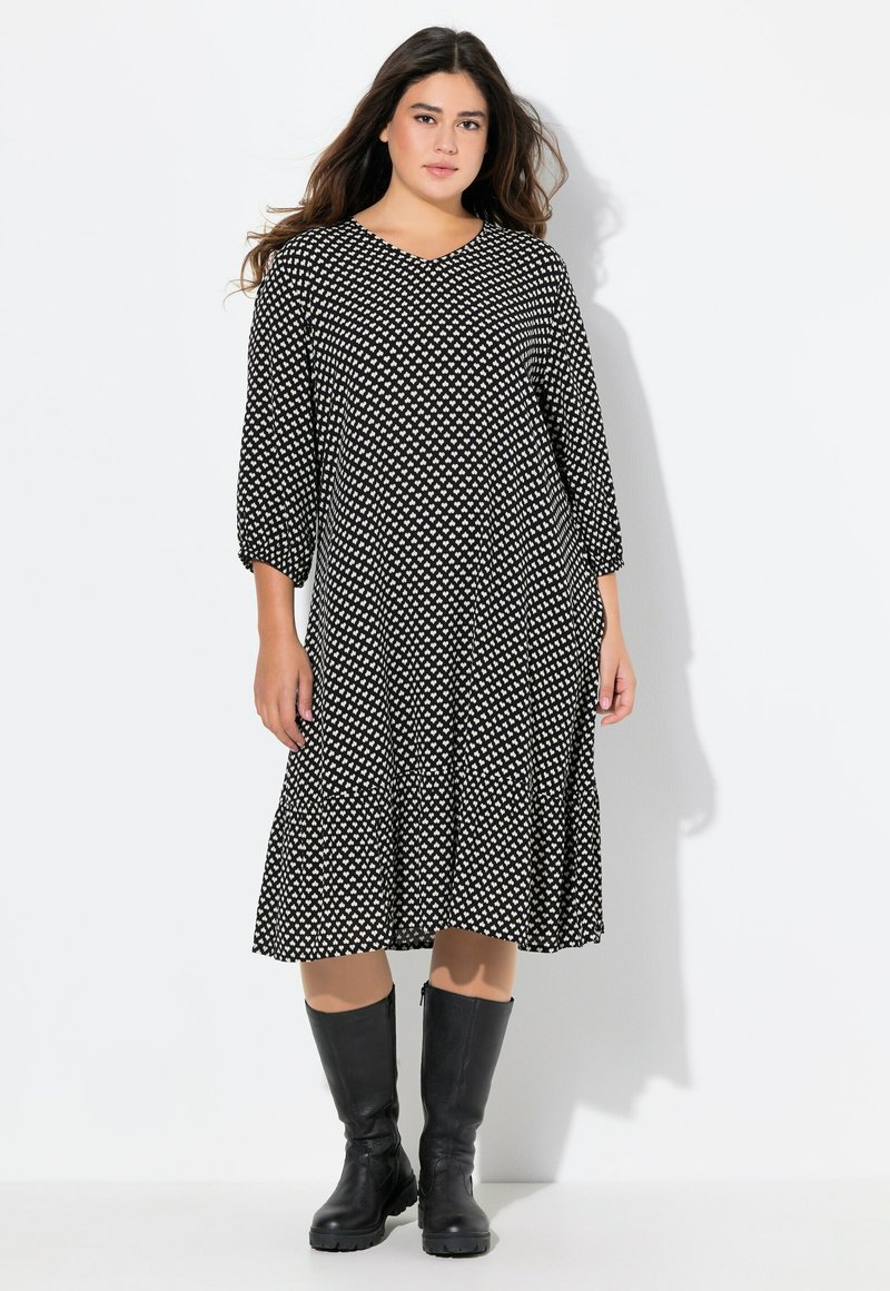 Schwarzes Kleid mit weißem Diamantmuster, V-Ausschnitt, Dreiviertelärmel und gestuftem Rock. Kombiniert mit knielangen schwarzen Stiefeln.