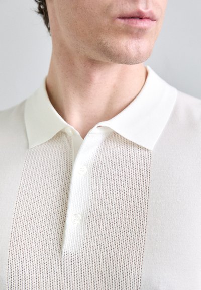 Ljusfärgad pikétröja med ett strukturerat stickat mönster i mitten, med en kontrasterande slät krage och tre knappar framtill.