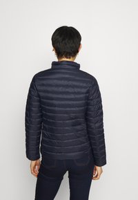 Person med kort mørkt hår iført en marineblå quiltet jakke og mørke bukser, set bagfra mod en ensartet lys baggrund.