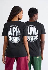 Melni T-krekli ar lielu baltu grafiku uz aizmugures, kurā rakstīts "ALPHA INDUSTRIES", ar brīvu piegriezumu un īsām piedurknēm.