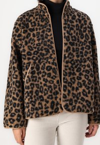 Leopardmönstrad, tan fleecjacka med svarta fläckar, som har en ståkrage, tryckknappar och två framfickor. Strukturerad tygfinish.