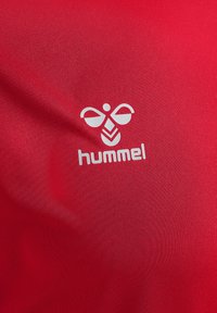 Röd sporttröja med en mjuk yta och en framträdande vit "hummel"-logotyp med en stiliserad grafik ovanför texten.