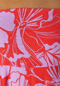 Tissu rouge avec un motif floral en violet clair, présentant de grands pétales et feuilles, mettant en valeur des couleurs vives et des détails de design complexes.