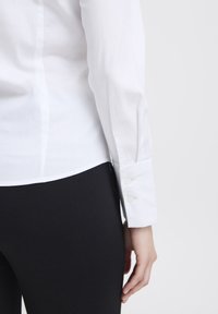 Camicia bianca con bottoni, dal tessuto liscio, forma aderente e maniche lunghe. Presenta un orlo arrotondato e polsini con un solo bottone. Indossata con pantaloni neri.