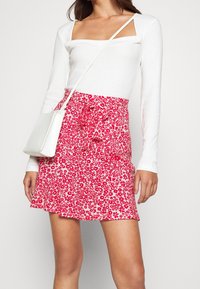 Haut blanc à manches longues avec décolleté carré, associé à une mini-jupe rouge à motifs floraux cintrée à la taille. Détail d'un sac bandoulière blanc visible.
