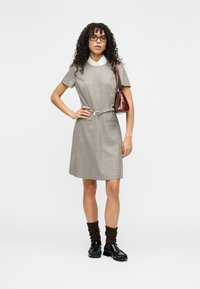 Καρό φόρεμα με κοντά μανίκια, γιακά και ζώνη. Φορεμένο με καφέ καλσόν μέχρι το γόνατο και μαύρα παπούτσια. Περιλαμβάνει μια μικρή καφέ τσάντα χειρός.