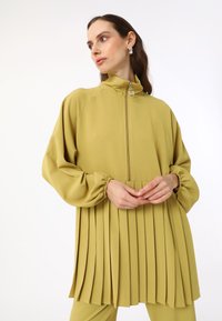 Heller olivgrüner, plissierter Blazer mit hohem Kragen, Reißverschluss und puffigen Ärmeln, aus einem glatten, strukturierten Stoff gefertigt.
