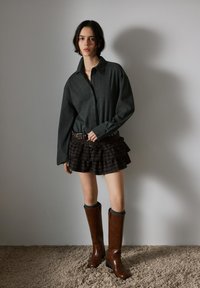 Jongere draagt een donker oversized overhemd, een gelaagde gerimpelde rok met riem en kniehoge bruine leren laarzen, staand op een tapijt.