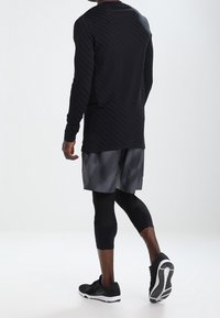 Musta pitkähihaine paita, jossa on tekstuurinen kuvio, yhdistettynä harmaisiin kuviollisiin shortseihin ja mustiin kompressiosukkiin, sekä mustiin lenkkikenkiin.