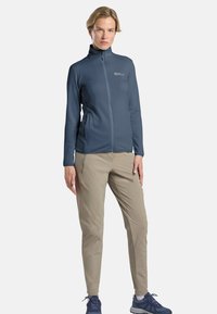 Veste zippée bleue avec un col montant, en tissu texturé, dotée de poches latérales, associée à un pantalon beige clair et des chaussures de sport bleues.