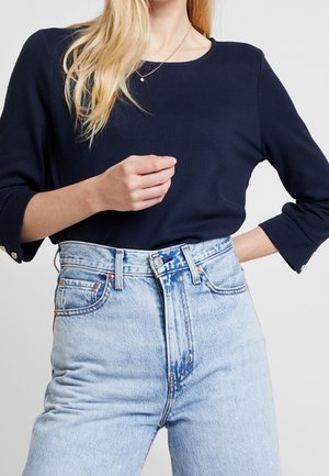 Femme portant un haut bleu marine à manches longues avec des détails de boutons et un jean en denim bleu clair taille haute, raccourci au niveau du cou et des hanches.