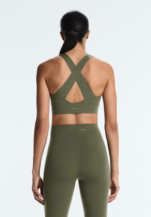 Sötét hajú nő lófarokkal, olívazöld keresztpántos sportmelltartót és hozzáillő, magas derekú leggingset viselve, háttal állva előrefelé.