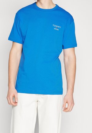 Man wearing a bright blue Tommy Jeans t-shirt and white pants, standing against a plain light background.