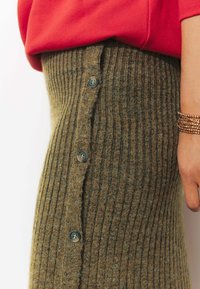 Jupe en tricot côtelé marron avec une fermeture latérale à boutons, comportant quatre boutons noirs. Un tissu texturé avec une coupe ajustée, associé à un haut rouge.