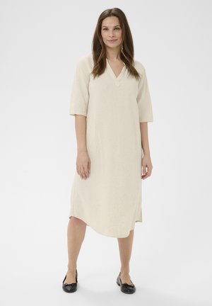 Kvinde stående iført en knælang cremefarvet kjole med albuelange ærmer og sorte balletsko mod en ensartet hvid baggrund.