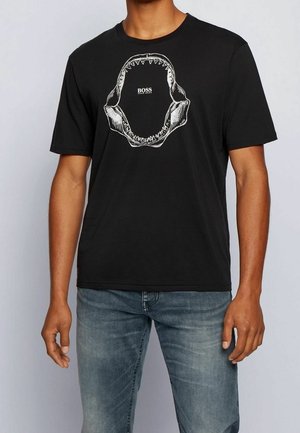 T-shirt en coton noir avec un graphique de mâchoire de requin en blanc. Manches courtes, col rond, coupe décontractée. Porté avec un jean bleu clair.