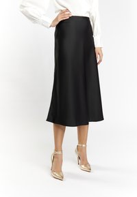 Schwarzer Satin-Midi-Rock mit glatter Textur und ausgestelltem Design. Getragen mit goldenen, spitzen Absätzen mit Knöchelriemen.