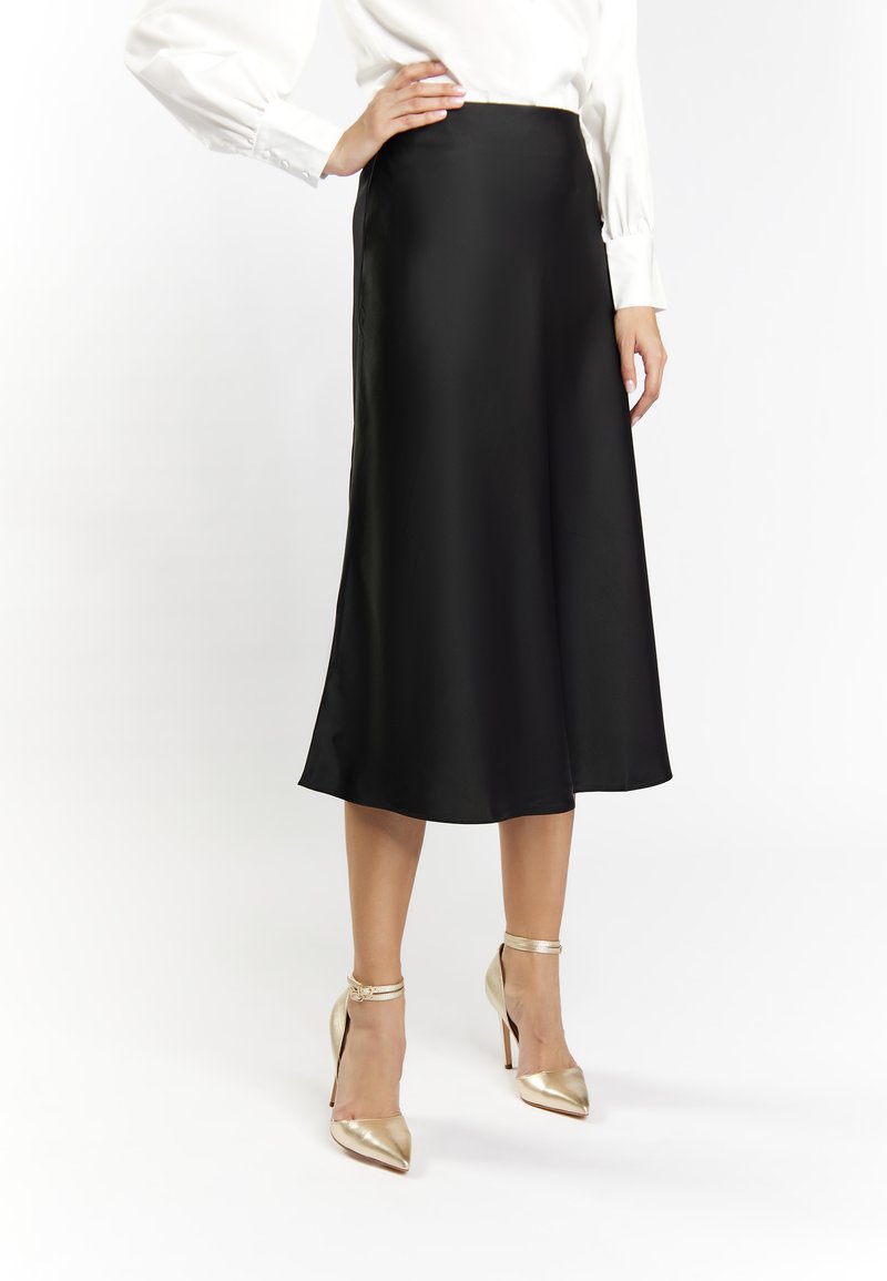 Schwarzer Satin-Midi-Rock mit glatter Textur und ausgestelltem Design. Getragen mit goldenen, spitzen Absätzen mit Knöchelriemen.