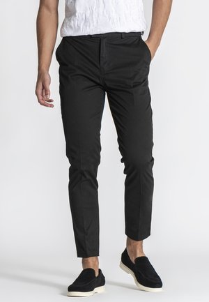 Homme portant un pantalon noir slim, une chemise blanche à manches courtes texturée et des chaussures noires à enfiler avec des semelles blanches, devant un fond uni.