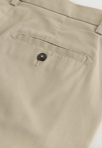 Primer plano de tela beige que muestra un bolsillo trasero en pantalones con un solo botón oscuro y detalles cosidos.