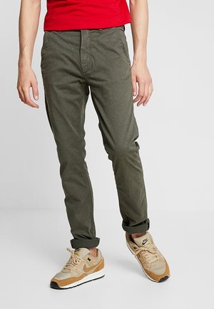 Hombre vistiendo pantalones ajustados verde oliva, zapatillas Nike beige y marrón claro, y una camiseta roja, de pie sobre un fondo gris.