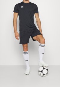 Miesurheilija pukeutuneena mustaan Under Armour -urheiluvaatteeseen ja valkoisiin Adidas-sukkiin, lepää jalka mustavalkoisella Adidas-jalkapallolla.