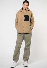 Beige Fleece-Hoodie mit schwarzem Taschen, Reißverschluss und Kordelzugkapuze. Kombiniert mit hellgrünen Hosen und weißen Turnschuhen.