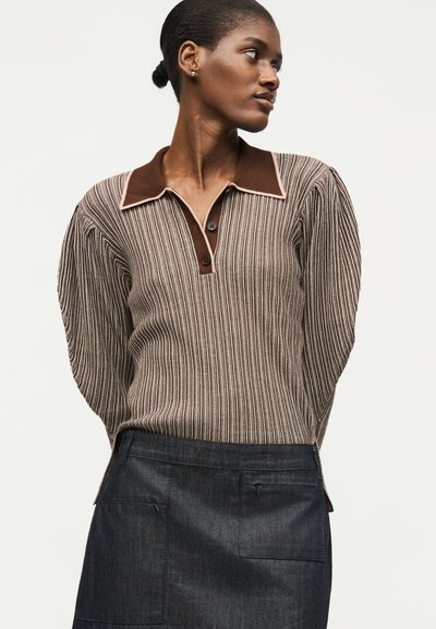 Γυναίκα με σκούρο δέρμα και μαλλιά μαζεμένα σε κότσο, που φοράει καφέ ριγέ πλεκτό τοπ με μακριά μανίκια, με σκουρόκαφε γιακά και σκουρόγκρι φούστα.