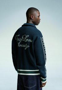 Jeune homme portant un pull bleu marine avec l'inscription blanche "West Lane Society" dans le dos, se tenant de profil contre un arrière-plan bleu-gris clair.