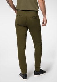 Slim-fit oliwkowe dresy wykonane z miękkiego materiału, z kieszeniami naszytymi z tyłu i prążkowanymi mankietami; noszone z czarnymi trampkami.