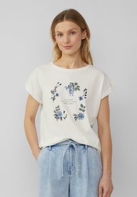 T-shirt en coton blanc cassé avec un col rond, des manches courtes roulées et un imprimé floral bleu et vert. Coupe décontractée, associé à un pantalon en denim bleu.