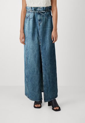 Maxinederdel i lyseblå denim med høj slids, høj talje og bindebælte. Modellen har kombineret den med sorte sandaler med blokhæle.