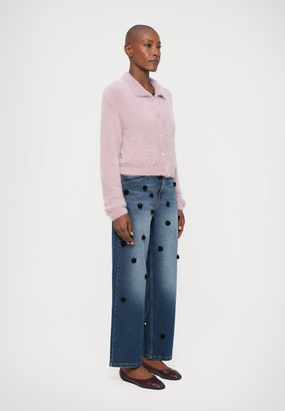 Svetlo ružový nadýchaný kardigán s golierom a gombíkmi, skombinovaný so širokými modrými džínami s čiernymi kvetinovými aplikáciami a postupným vyblednutím.