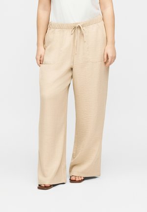 Pantalon beige à jambes larges avec taille élastique et cordon, poches plaquées devant, porté avec des sandales marron et un haut blanc.