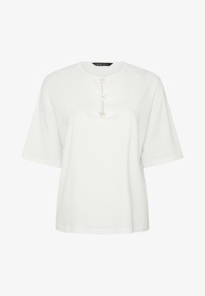 Top bianco a maniche corte ampie con scollo rotondo e tre bottoni decorativi a forma di fiore bianco lungo il centro anteriore.