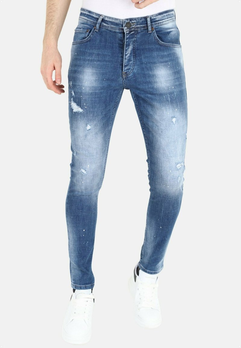 Slanke blauwe spijkerbroek met beschadigde details, vervaagde delen en lichtblauwe accenten. Gecombineerd met witte sneakers.