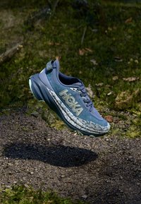 Chaussure de course légère de couleur bleue avec une tige texturée, un logo HOKA bien visible et une semelle rembourrée arborant un motif blanc et gris.
