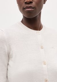 Prim-plan cu o persoană purtând un cardigan bej deschis cu nasturi rotunzi și un logo subtil brodat pe piept.