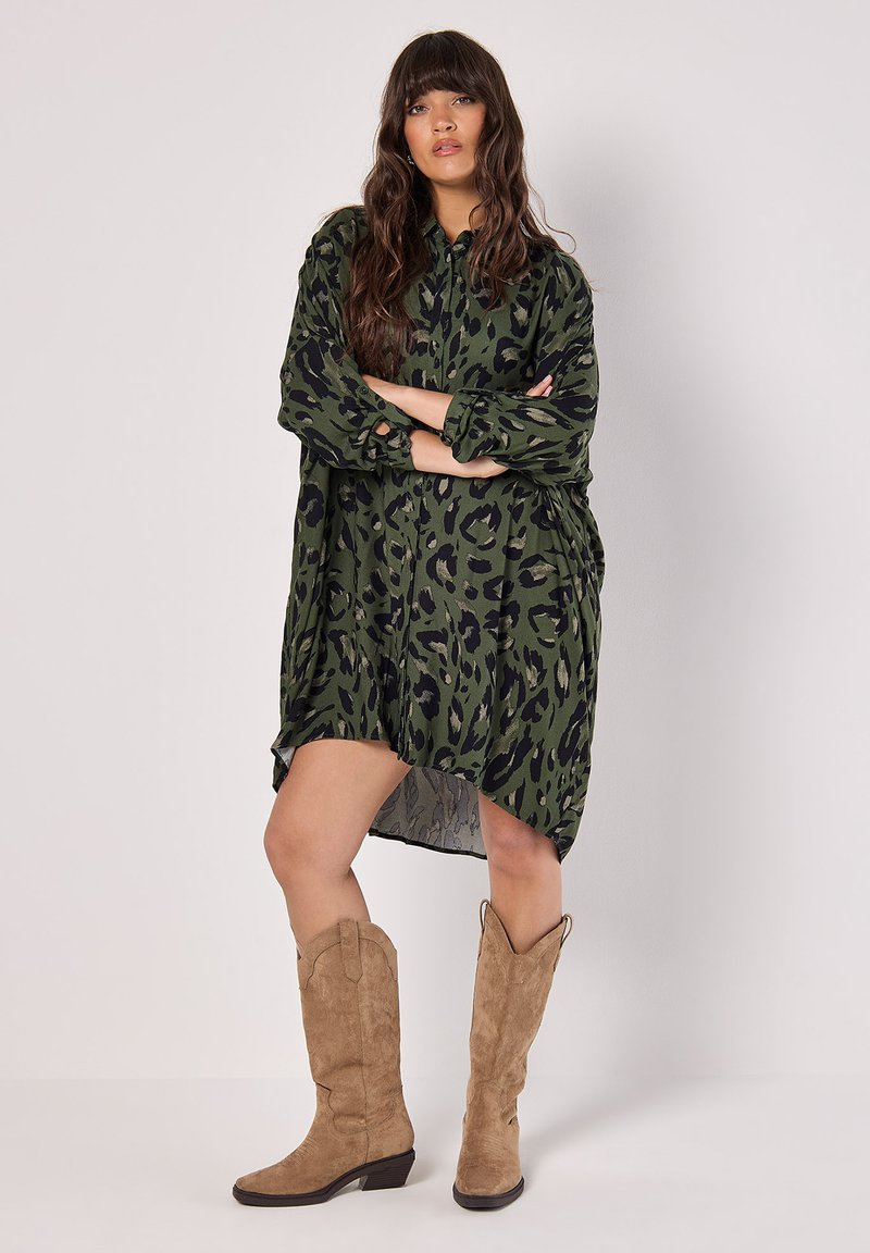 Vestido camisero de estampado de leopardo verde hecho de tela ligera, con un frente de botones, mangas largas y un dobladillo asimétrico. Combinado con botas de ante beige.