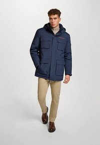 Marineblauwe geïsoleerde parka met capuchon, meerdere voorzakken en ritssluiting. Draagt over een lichtgrijze trui en beige broek.