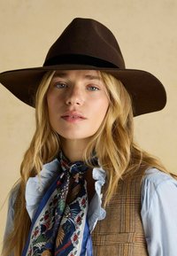 Pălărie maro din pâslă cu boruri largi, combinată cu o cămașă albastră cu guler volan și un fular decorativ albastru legat la gât.