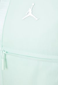 Gaiši piparmētru zaļa mugursoma ar teksturētu audumu, kas aprīkota ar rāvējslēdzēja kabatu un baltu Jumpman logo, kas skaidri redzams priekšpusē.