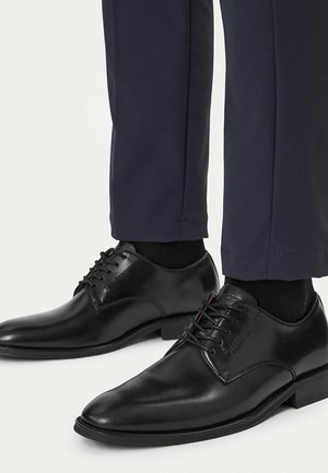 Schwarze, polierte Leder-Schnürschuhe, getragen mit schwarzen Socken und dunkelblauen, maßgeschneiderten Hosen vor weißem Hintergrund.